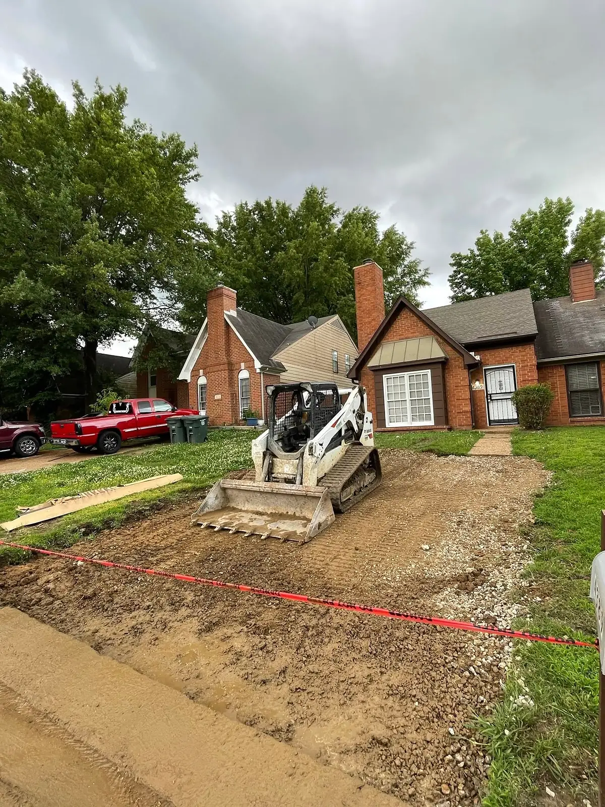 Skid steer grading the base for a new driveway beside a brick home